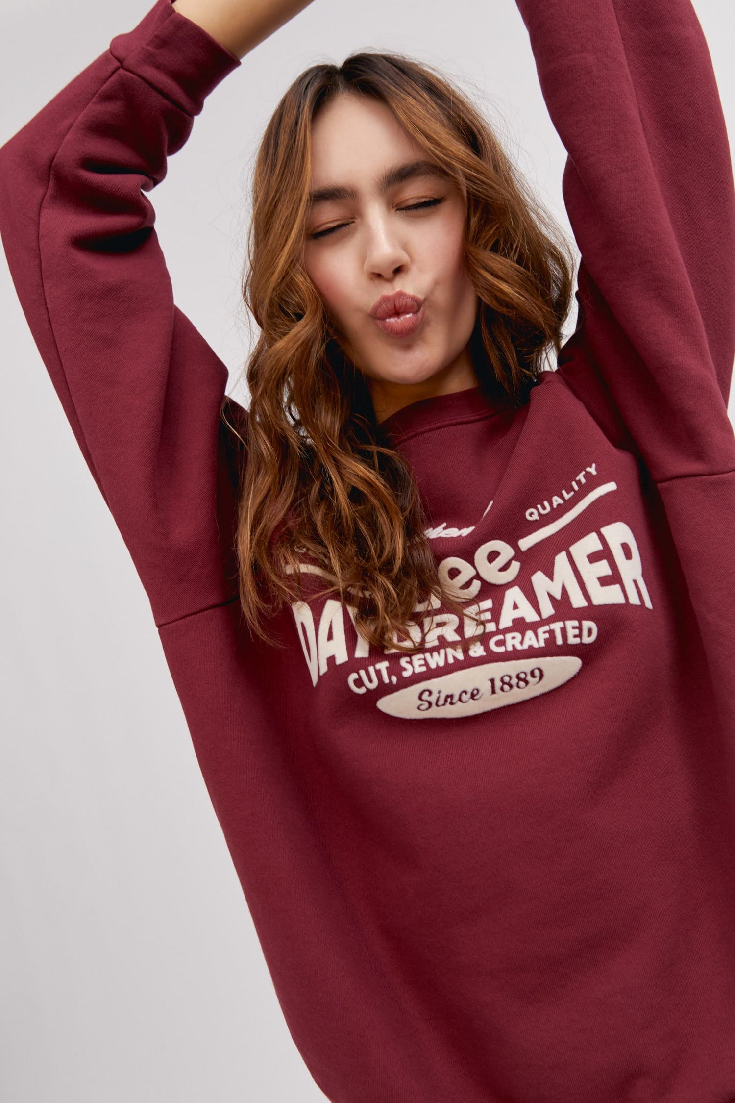 long haired model making kissing face and wearing maroon colored sweatshirt with flocked logo graphics
