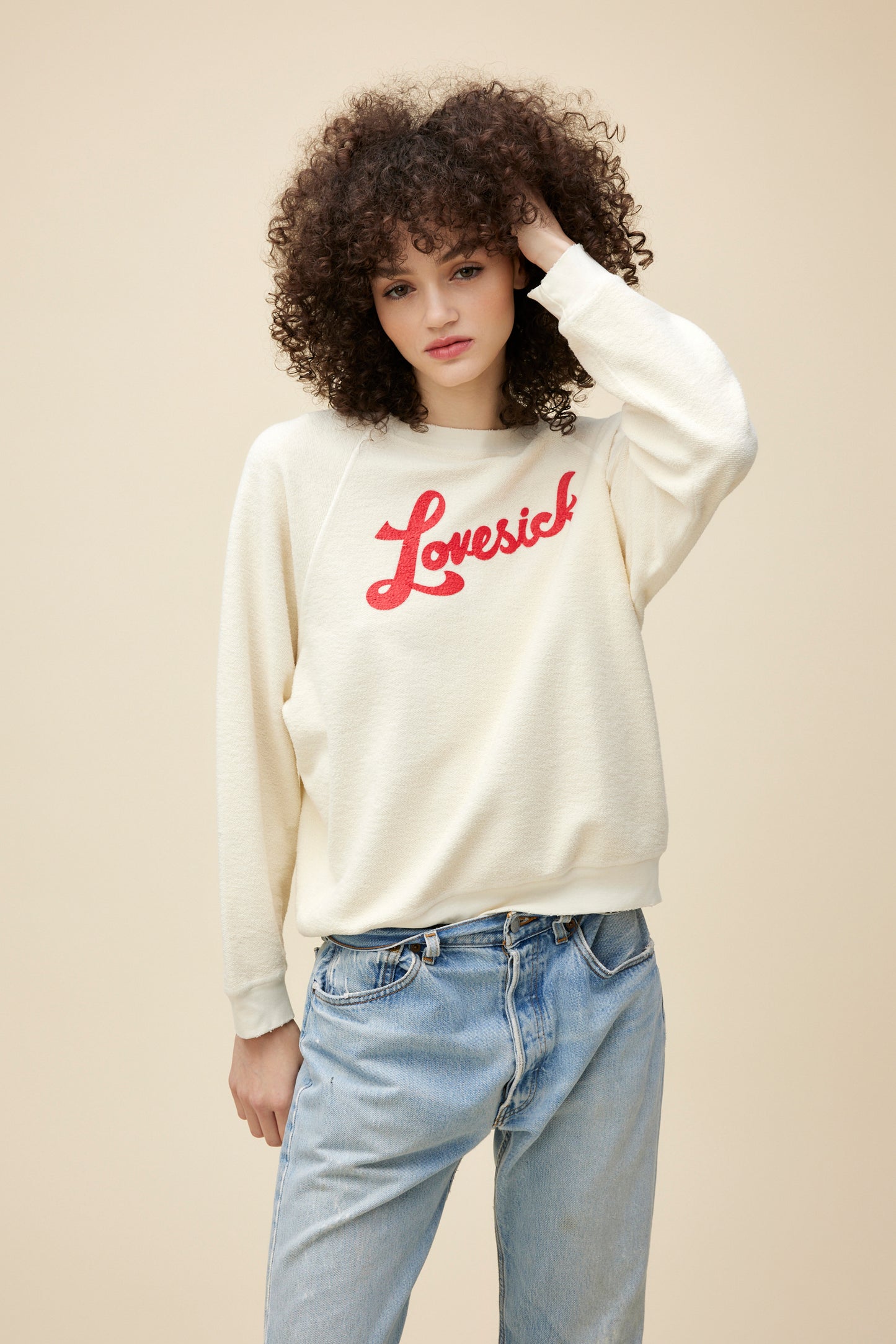 A curly-haired model wearing a reverse brushed fleece sweatshirt stamped with 'lovesick' red script on the front.