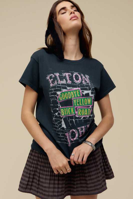 A model featuring a vintage black tee featuring motifs from Elton John's Goodbye, Yellow Brick Road album's original cover, designed with a brick road graphic and accented with rhinestones.