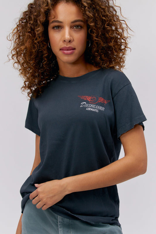 A curly-haired model featuring a black tee stamped with 'Daydreamer Airwaves' and the daydreamer logo with wings front and back.
