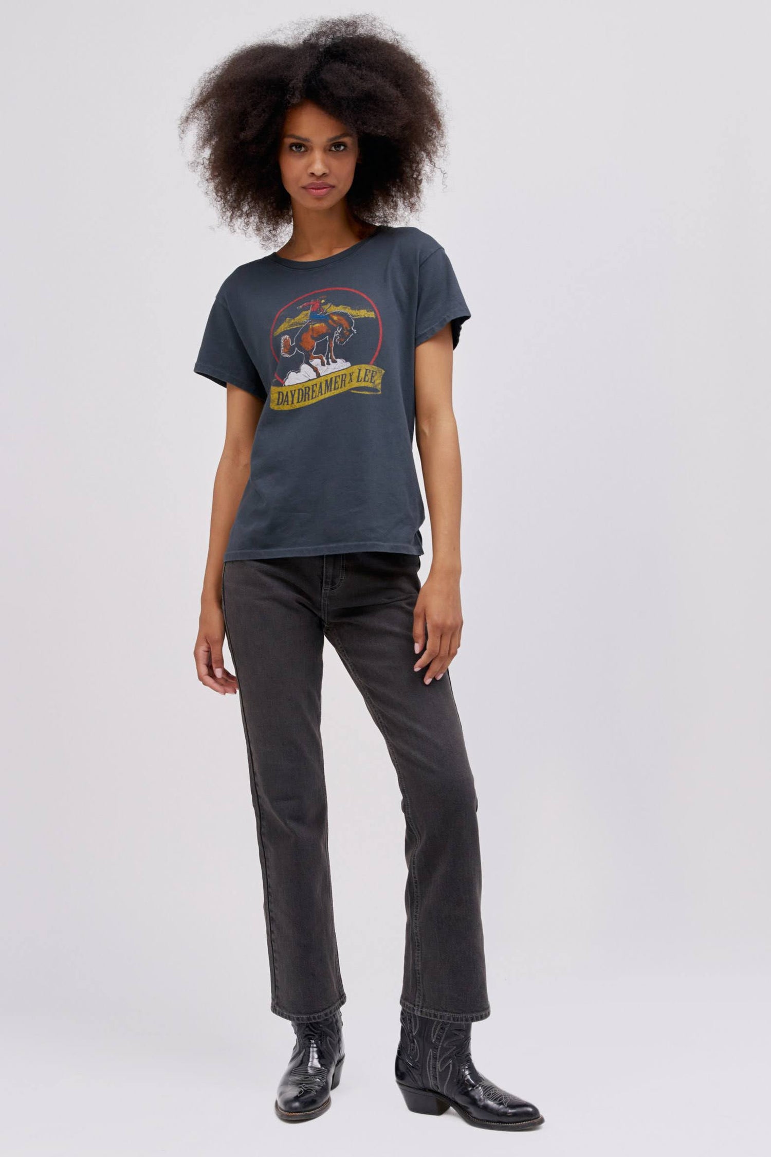 A curly-haired model featuring a black tour tee designed with a fresh rendition of a tried and true Lee vintage rodeo graphic hits a relaxed fit, Tour Tee.