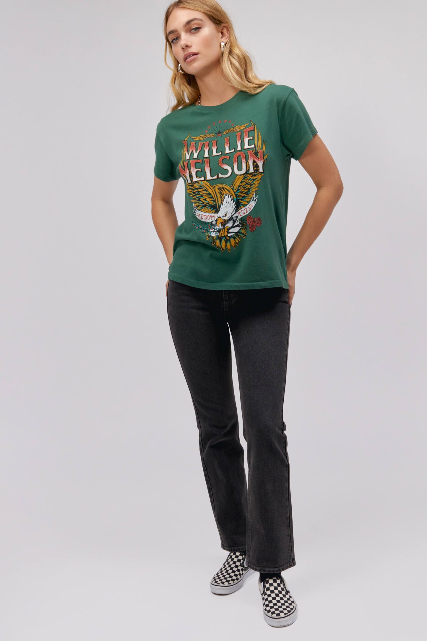 A blonde-haired model featuring a green tee stamped with 'Willie Nelson' and designed with a bird with 'Abott Texas' on its mouth.