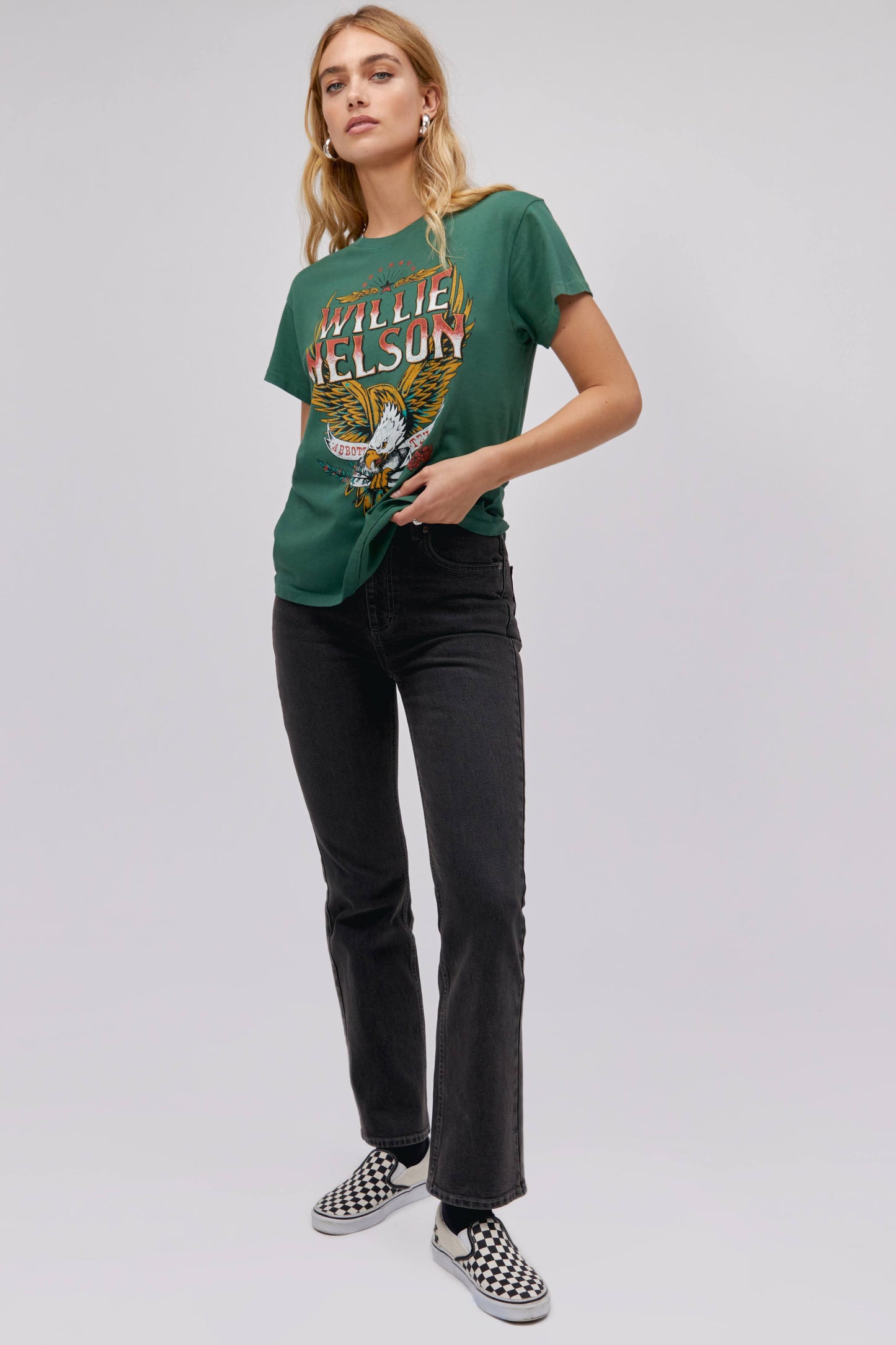 A blonde-haired model featuring a green tee stamped with 'Willie Nelson' and designed with a bird with 'Abott Texas' on its mouth.