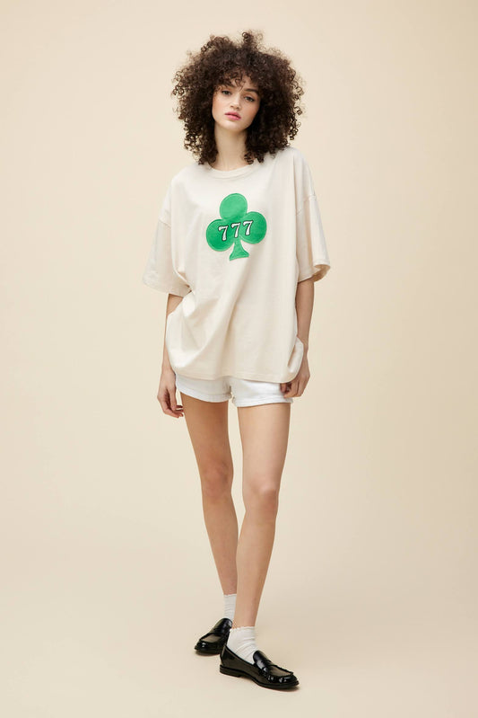 Curly-haired model wearing an oversized tee with a patched clover detail and 777 graphic.