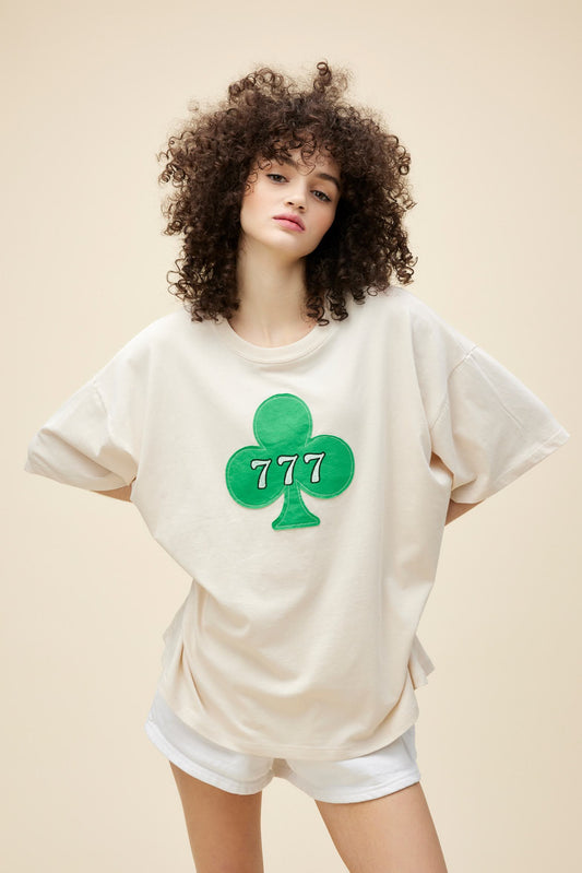 Curly-haired model wearing an oversized tee with a patched clover detail and 777 graphic.