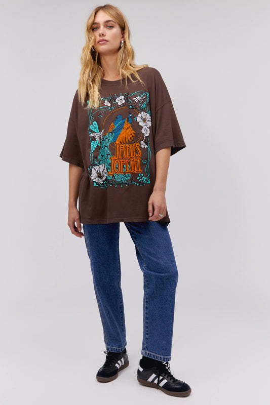 A blonde-haired model featuring a dark brown tee stamped with Janis Joplin in orange, designed with a peacock, leaves, and flowers around, accented in orange, blue, and white.