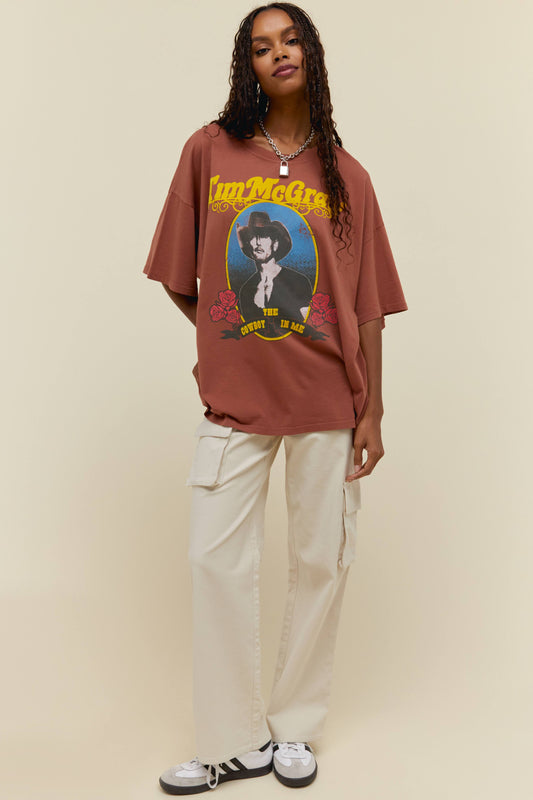 A dark-haired model featuring a reddish brown t-shirt dress adorned with Tim McGraw" on top with the graphic image of Tim Mcgraw in the center