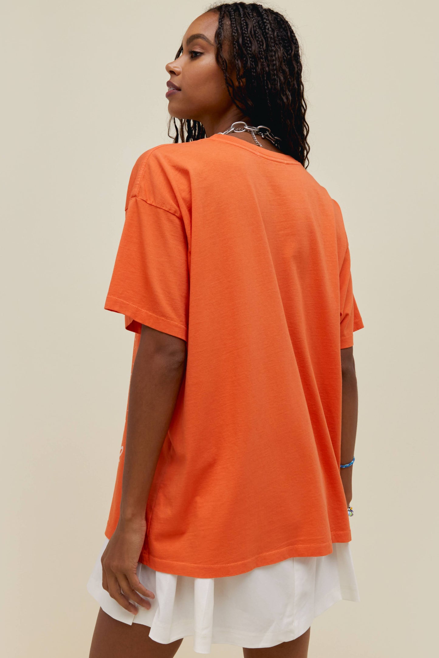 A dark-haired model featuring an orange tee stamped with "Elton John" on the upper part of the tshirt centered with a graphic image of Elton John on his 1977 tour