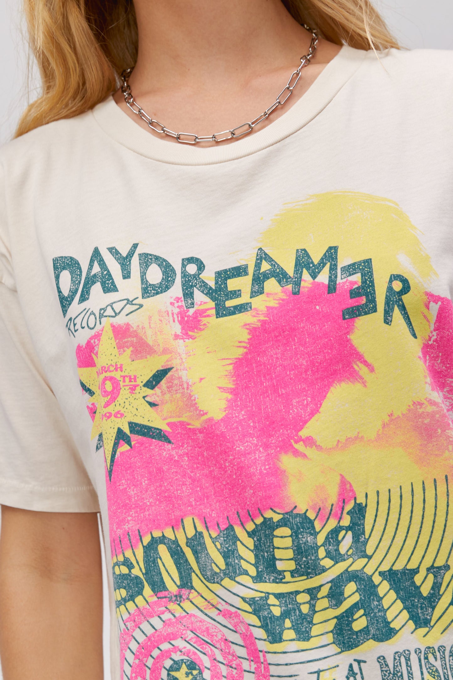 A blonde-haired model featuring a white tee stamped with 'daydreamer' and 'sound waves' at the back. It is also designed with pink and yellow graphics.