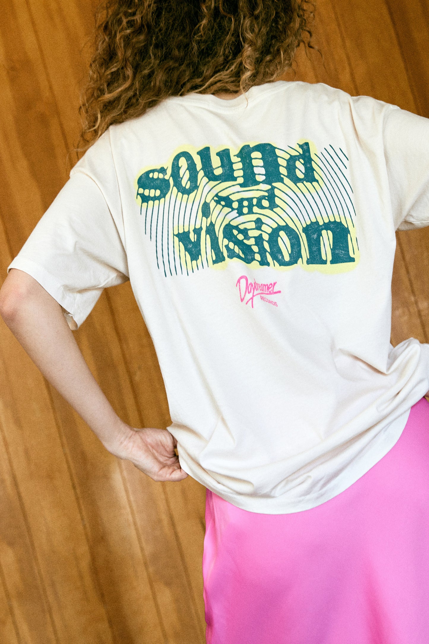 A curly-haired model featuring a white tee stamped with 'daydreamer' and 'sound waves' at the back. It is also designed with pink and yellow graphics.