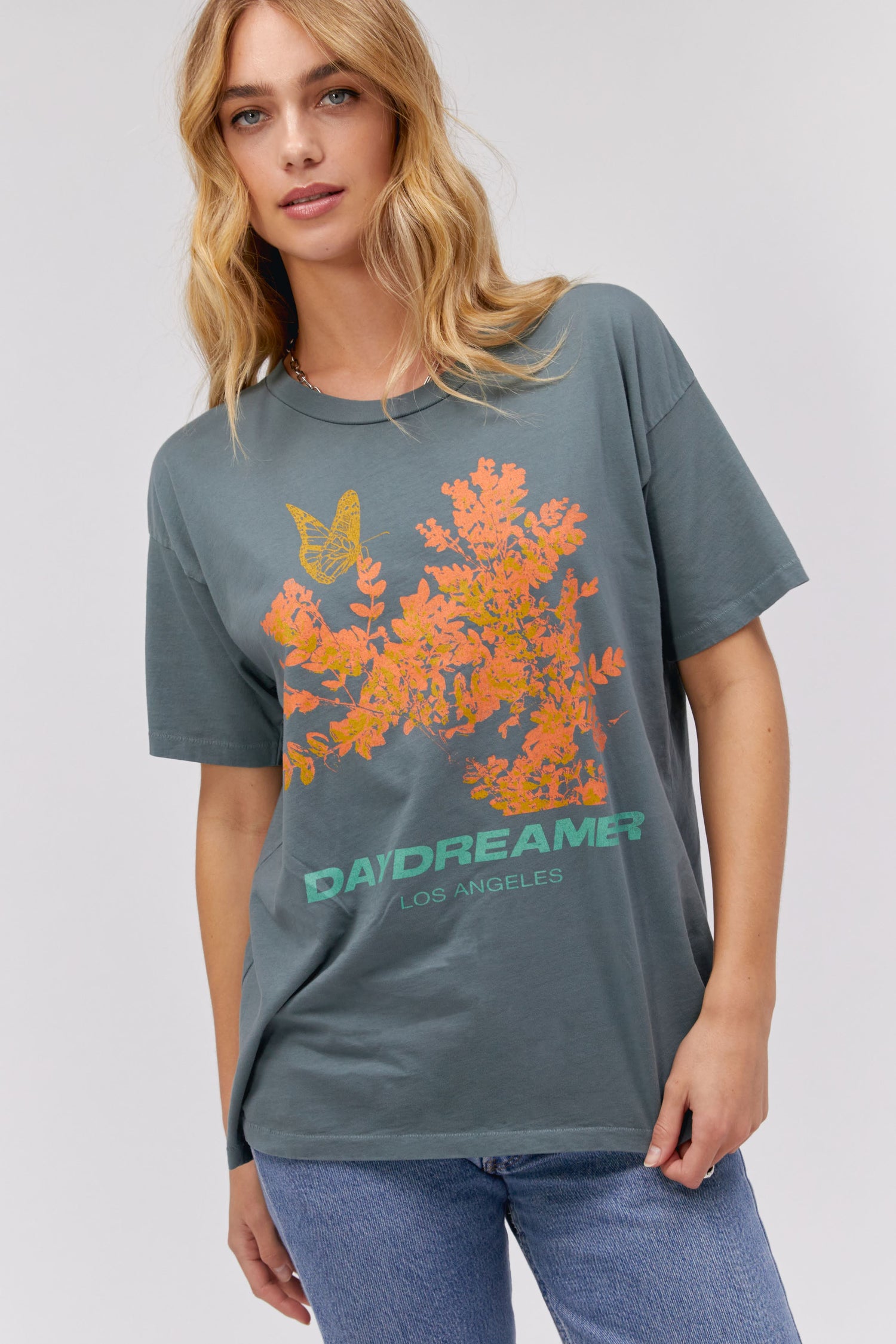 A blonde-haired model featuring a grey tee stamped with 'Daydreamer Los Angeles', a graphic of leaves and butterflies in orange, the same at the back but stamped with 'Bloom'.