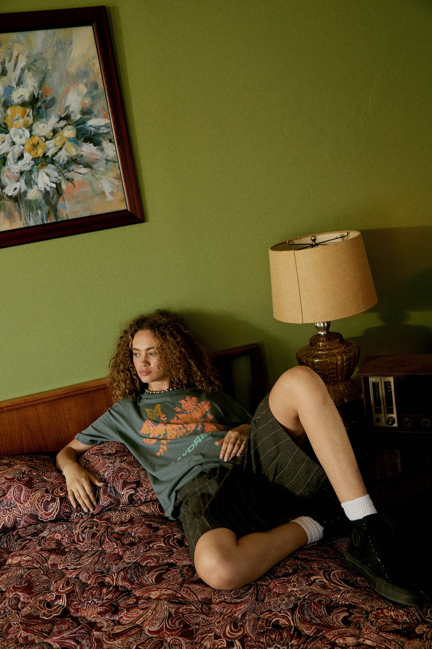 A curly-haired model featuring a grey tee stamped with 'Daydreamer Los Angeles', a graphic of leaves and butterflies in orange, the same at the back but stamped with 'Bloom'.