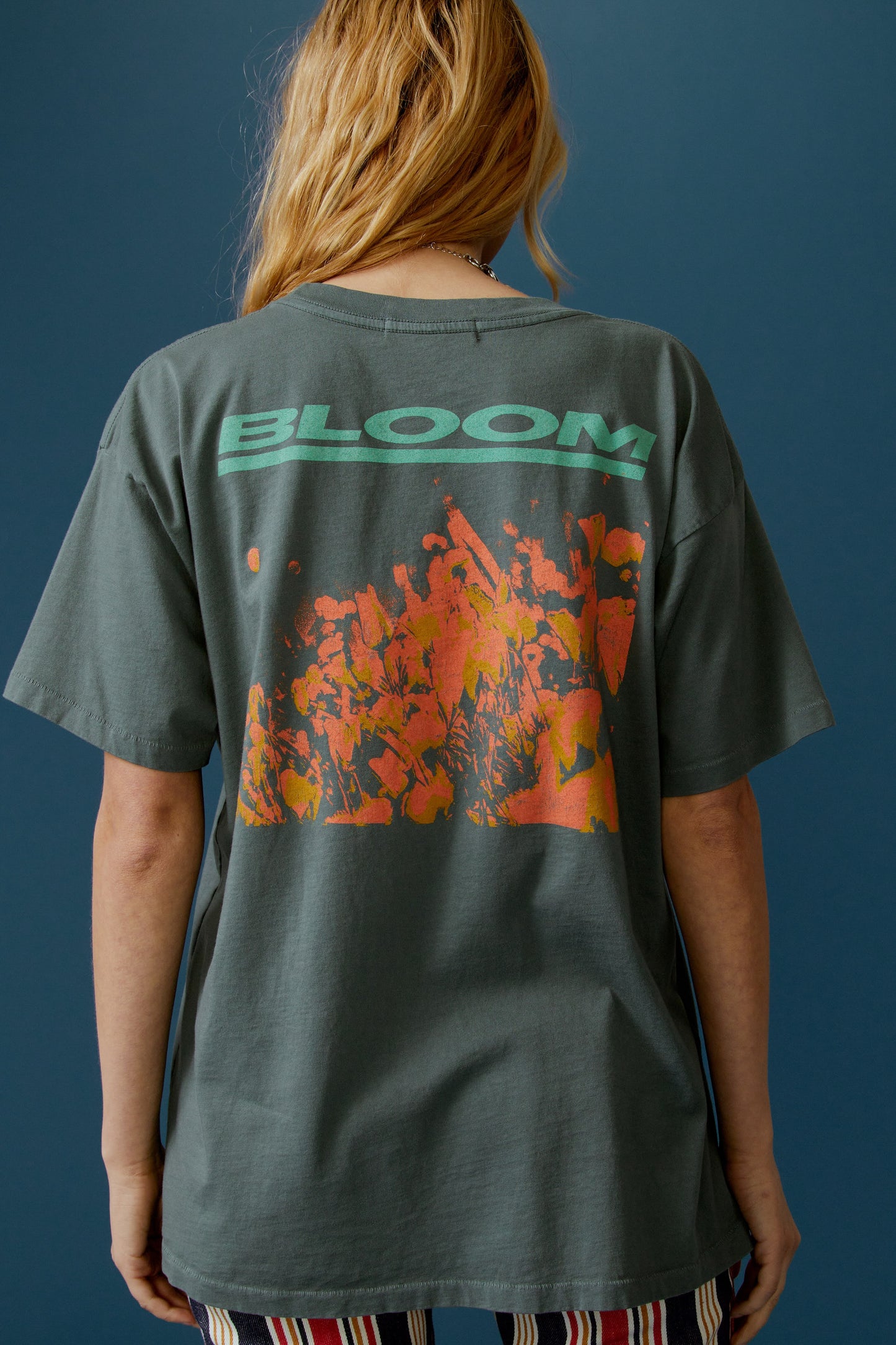 A blonde-haired model featuring a grey tee stamped with 'Daydreamer Los Angeles', a graphic of leaves and butterflies in orange, the same at the back but stamped with 'Bloom'.