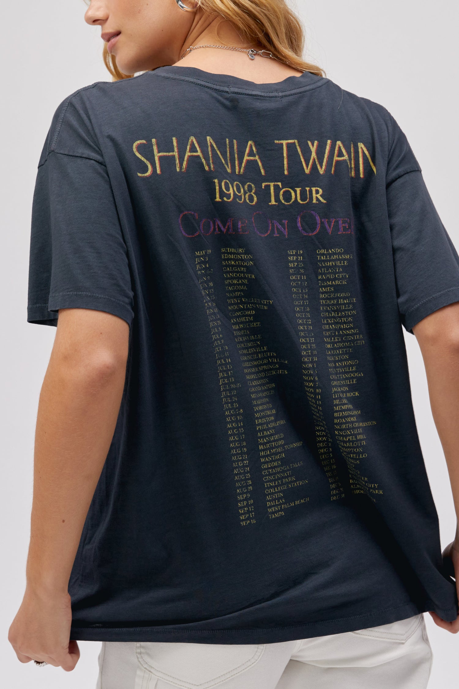 A blonde-haired model wearing a black merch tee featuring an iconic portrait of the female artist on the front and the list of her tour dates and cities at the back.