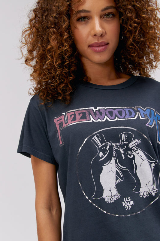 A curly-haired model featuring a black tee stamped with 'Flletwood Mac' and designed with penguin artwork in black and white.
