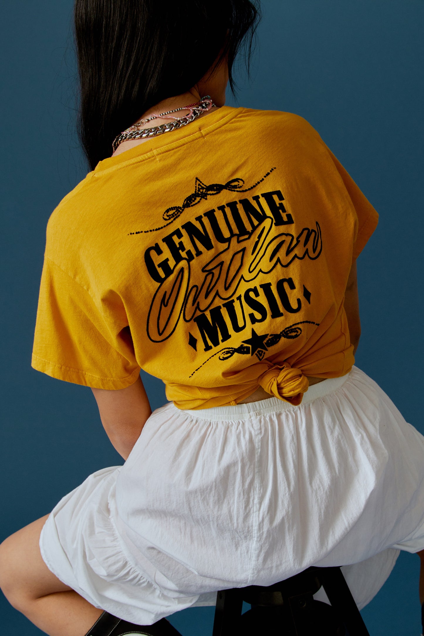 A dark-haired model featuring a golden daze tee stamped with 'Willie Nelson' in front and 'Genuine Outlaw Music' at the back.