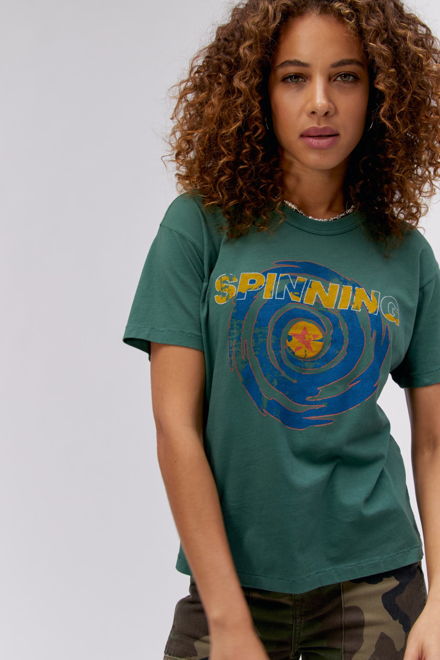 A curly-haired model featuring a green tee stamped with 'Spinning' and a spinning whirl with a star in the middle.