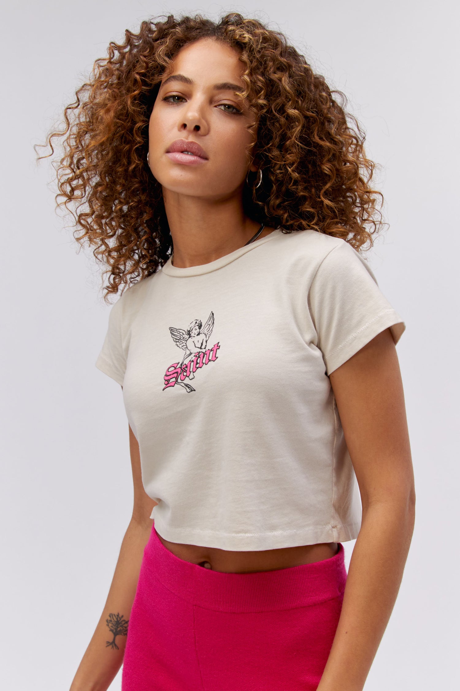 A curly-haired model features a white cropped tee designed with a baby angel in the center, stamped with 'Saint' in pink font in front.