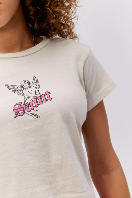 A curly-haired model features a white cropped tee designed with a baby angel in the center, stamped with 'Saint' in pink font in front.