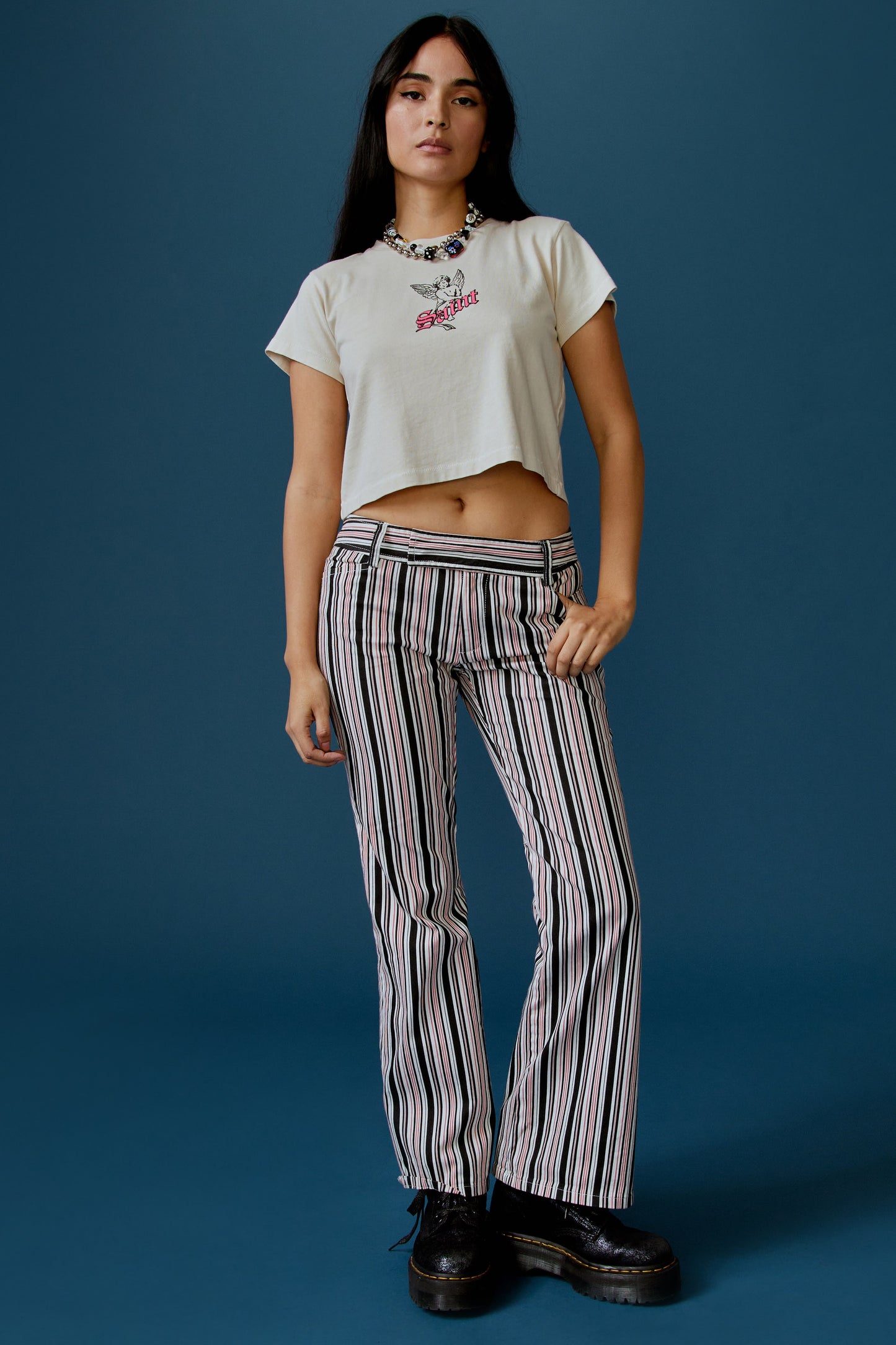 A dark-haired model features a white cropped tee designed with a baby angel in the center, stamped with 'Saint' in pink font in front.