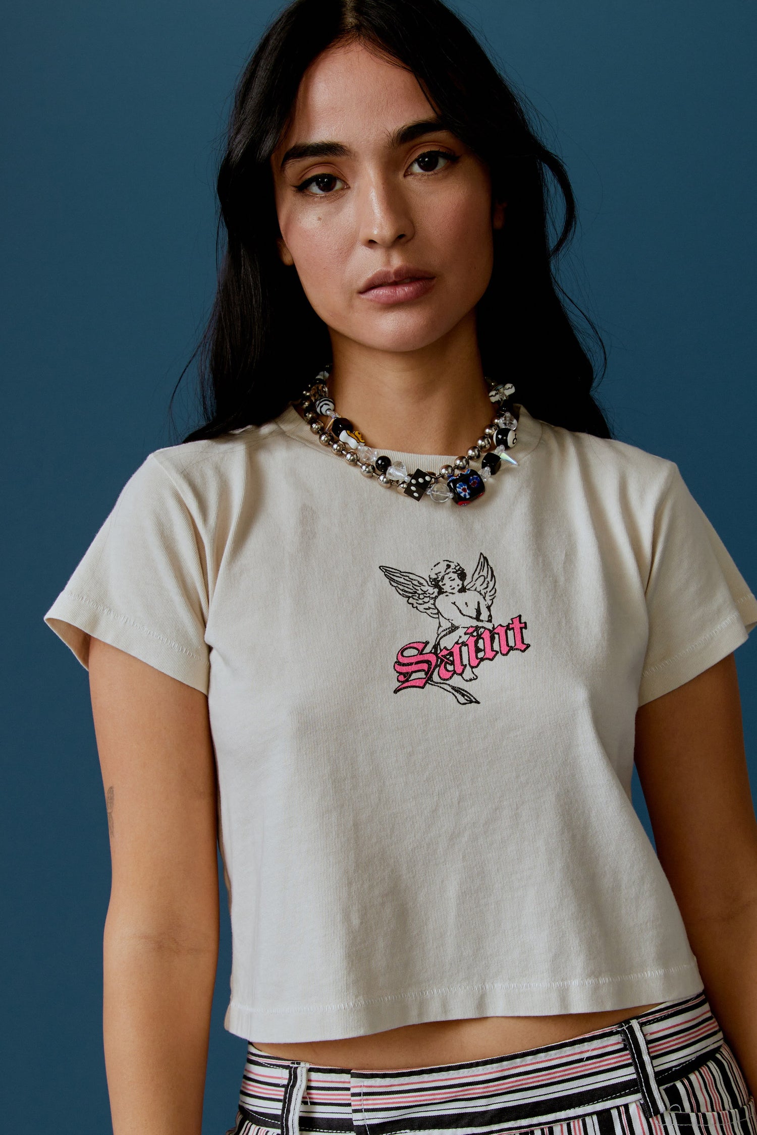 A dark-haired model features a white cropped tee designed with a baby angel in the center, stamped with 'Saint' in pink font in front.
