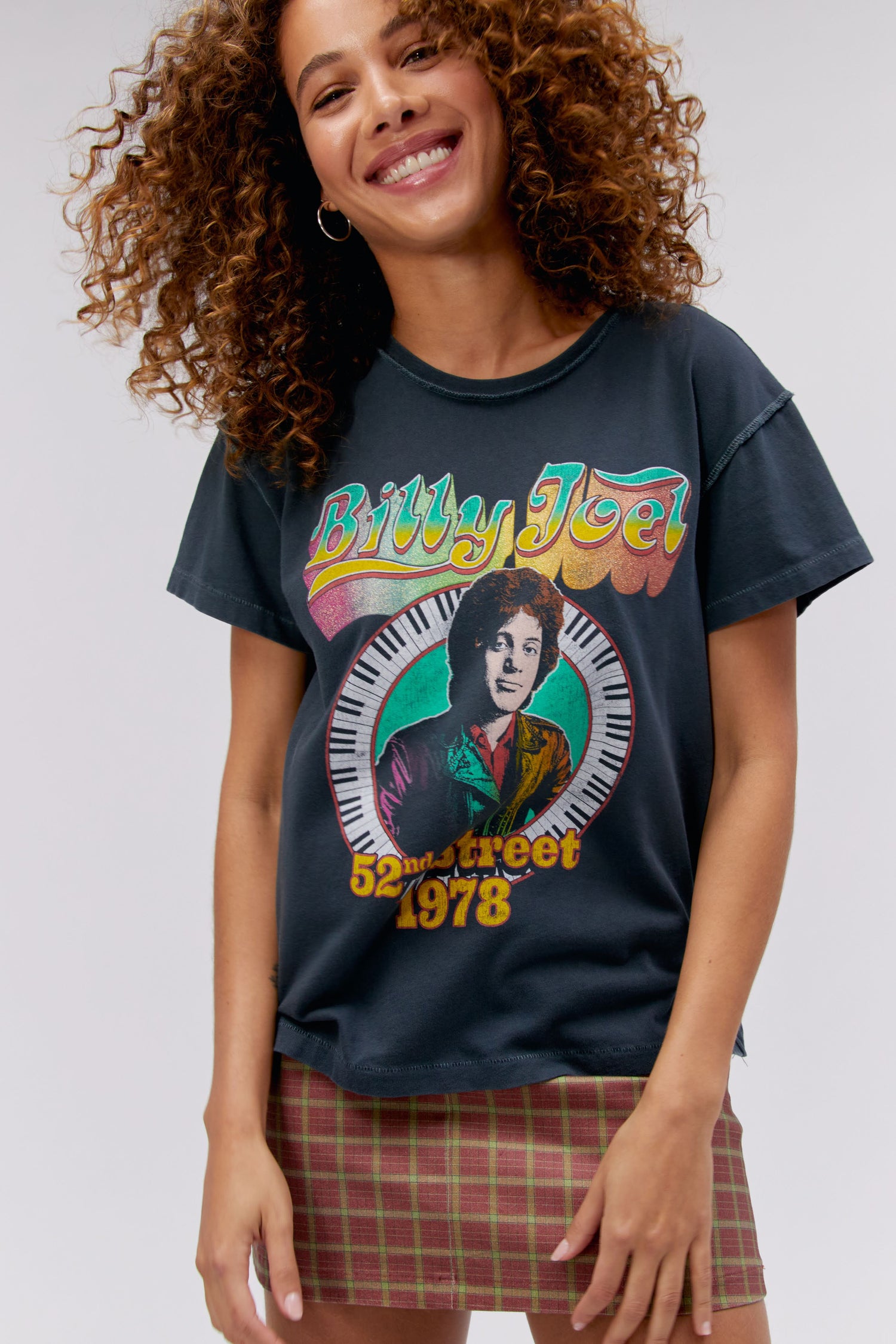 A curly-haired model featuring a black tee stamped with 'Billy Joel' in blue, green, and yellow gradient letters, along with a portrait of the artist in the center.
