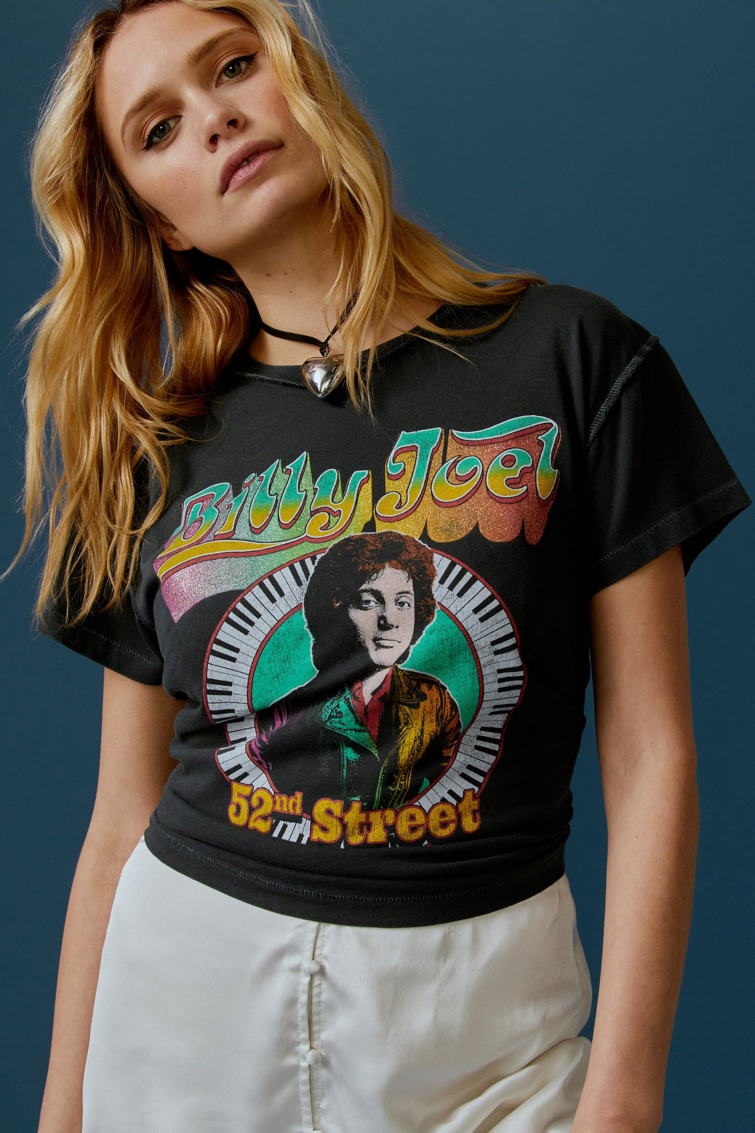 A blonde-haired model featuring a black tee stamped with 'Billy Joel' in blue, green, and yellow gradient letters, along with a portrait of the artist in the center.