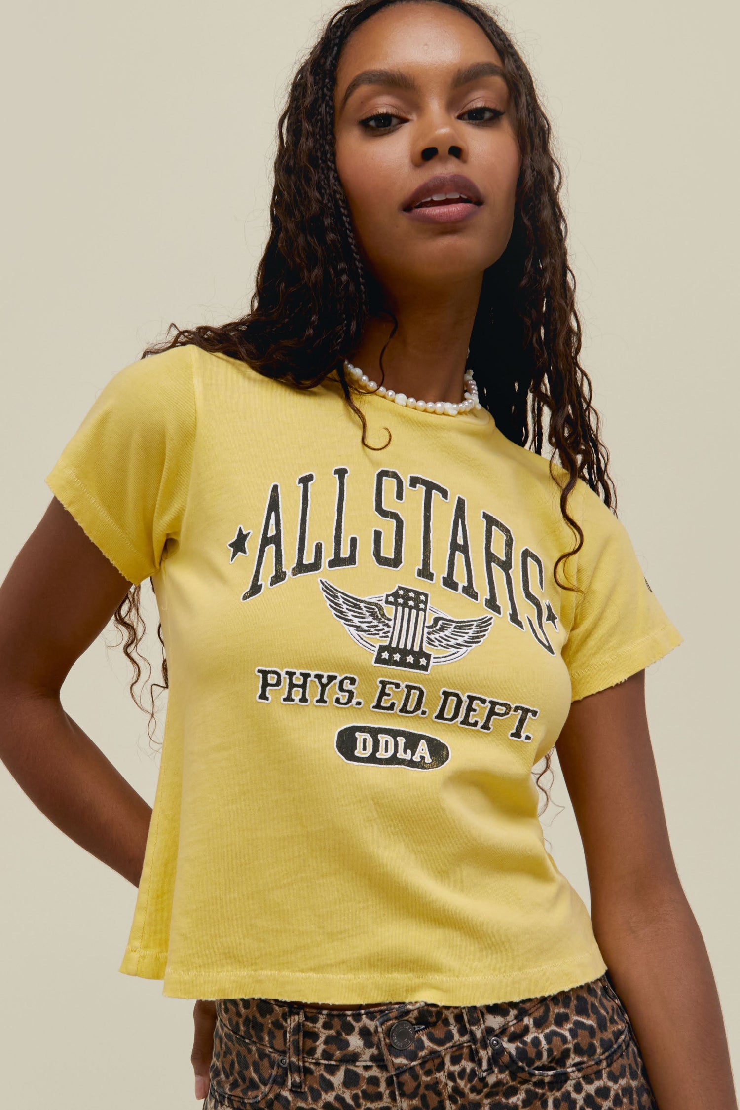 A dark-haired model wearing a yellow cropped tee featuring a large font "ALL STARS PHYS ED DEPT" and a graphic logo