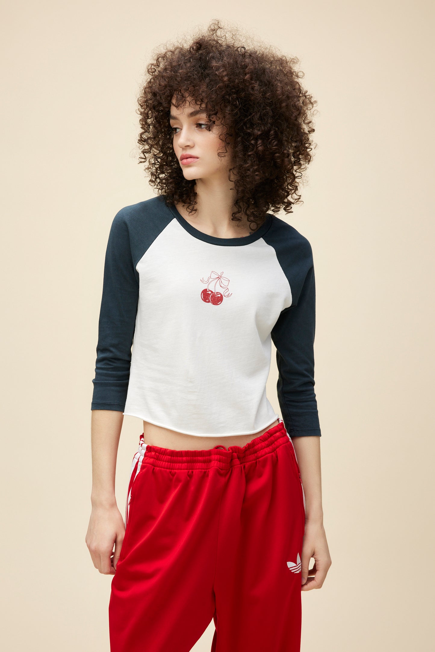 A curly-haired model wears a vintage-inspired long sleeve raglan tee with a red cherry graphic on the front.