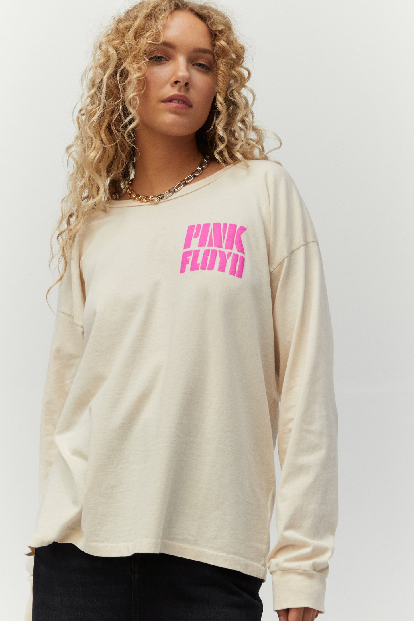Blonde curly-haired model featuring a white-colored long sleeve designed with psychedelic-inspired block letters and a stamp of Pink Floyd’s inflatable pig, Algie, accented in pink and green puff ink
