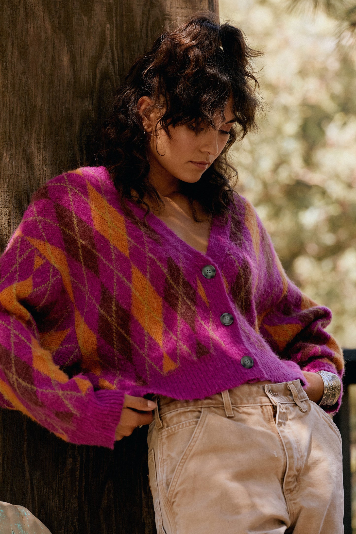 Dark curly-haired model featuring  a relaxed, slightly cropped, ultra cozy cardigan designed in a standout argyle pattern in unexpected color combos of fuschia, orange and gold