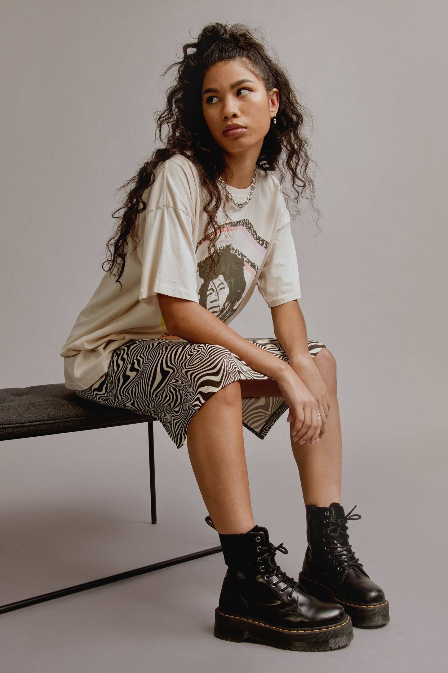 Dark curly-haired model featuring a dirty white tee designed with a multi-colored spiral with the icon’s portrait and name in hand-drawn letters